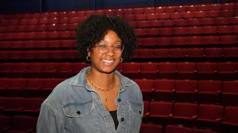 Shaun Whitmore/BBC Becki Blackwood standing in the New Wolsey Theatre. Behind her are rows of empty seats. The seats are a rich red colour. Becki is wearing a blue denim shirt, a gold necklace and is wearing glasses. 