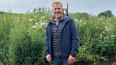 BBC Adam Henson, wearing all navy blue - blue jeans and a blue waterproof - stands in front of a tall bed of wildflowers