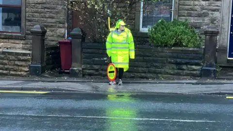 A woman stands on the other side of the road, looking to one side. She is wearing a fluorescent yellow coat and is holding a stop sign. Behind her there are houses.