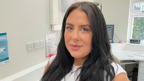 Wearing a white sleeveless top and with long dark brown hair, Beth looks into the camera. She also has glasses resting on her head and a nose ring in her left nostril. She stands inside a white walled room, with a window partially visible behind her. 