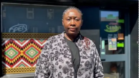 Shows a woman with her hair in plaits and a cardigan standing in front of a restaurant 