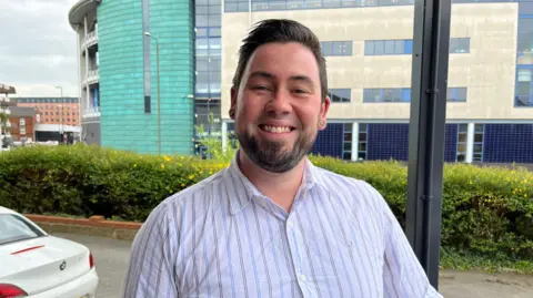 Man with dark hair and beard smiling in lilac striped shirt 