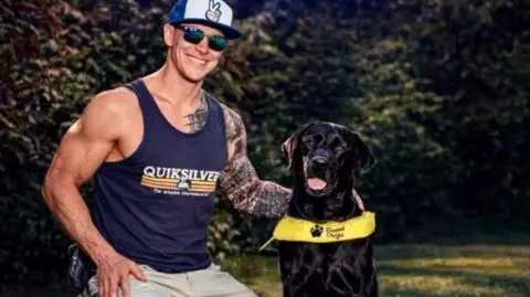 Mark Long Wearing a blue baseball cap and top with shorts. He is knelt down beside his black Labrador guide dog Mary.