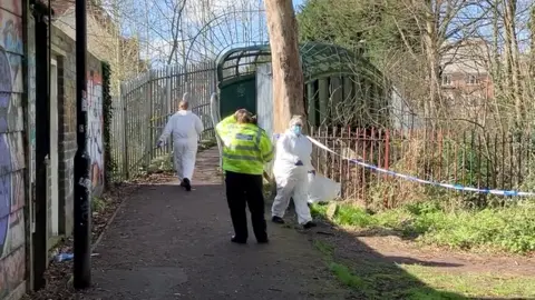 Two white-suited forensics officers can be seen walking in the park. A female police officer wearing a high-vis jacket is watching them. 