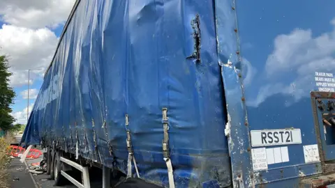 View of the side of the large HGV trailer, covered in blue plastic, with a large bulge