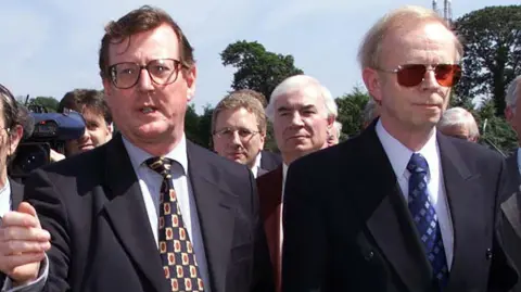 Pacemaker David Trimble on left of picture in a navy suit gesturing towards the camera. He has brown hair and glasses. 
Lord Empey on the right is also in a navy suit. He has blonde hair and is wearing sunglasses