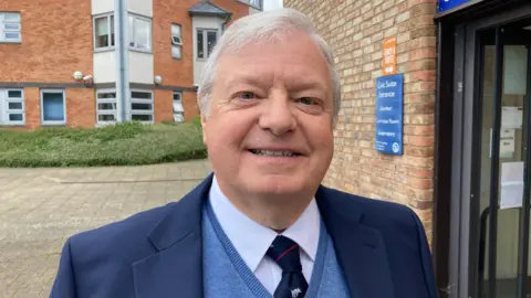 A man standing outside a bricked building, wearing a blue suit, blue v-necked jumper and a navy and red tie. 