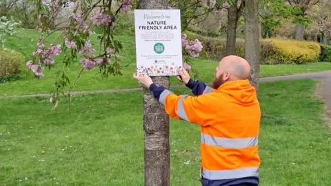 A bald man with a brown beard wearing an orange hoody attaches a white sign to a tree.