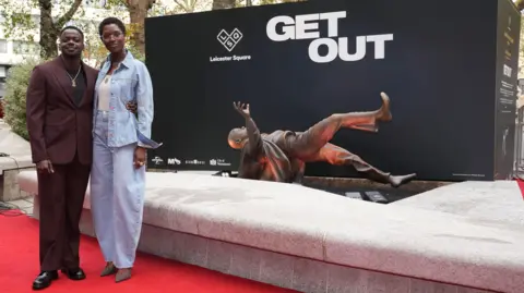 PA Images Jodie Turner-Smith and Daniel Kaluuya pose on a red carpet beside the horizontal statue of Mr Kaluuya as his character in Get Out. Mr Kaluuya is dressed in a mauve suit and Ms Turner-Smith is wearing a blue denim jacket and trousers. 