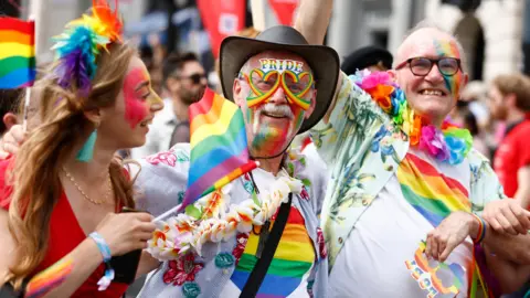 Getty Images Two men and a woman at a pride march. They are all wearing rainbow clothing and face paint. The two men are looking ahead and smiling while the woman next to them is looking to the side of them.
