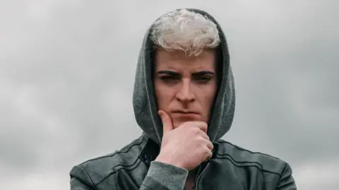 A serious-looking young man stands against a blurred background, his hand placed thoughtfully on his chin. His eyebrows are darkened, his hair a shock of white on top of his head, peeking out from a grey hood. He wears a dark grey leather jacket.