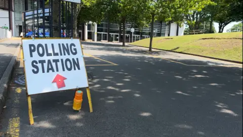 BBC A sign which says polling station with a red arrow pointing towards Beau Sejour, a leisure centre with trees outside and some greenery on the right. 