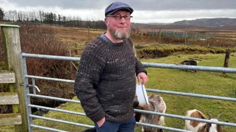 A man wearing a made -made man stands in front of a gate holding a bowl with small circular glasses and a beard. 