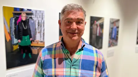 Rob Jones, CEO of Les Bourgs stood in front of four of the portraits. He is smiling and wearing a multi-coloured shirt.