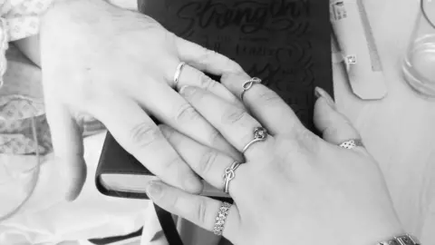 Rachel Lindley A black and white image of two hands interlocking. The man's hand on the left has a wedding ring on and the woman's hand has five rings on, one of which is a wedding ring.