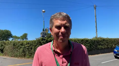 Man in front of a crossroad behind him is a bush and a lamp post. He is wearing a pink polo shirt with a green lanyard. 