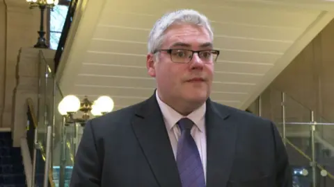 Gavin Robinson wears a purple tie, off white shirt, and black suit. He has grey hair and dark rimmed glasses. 