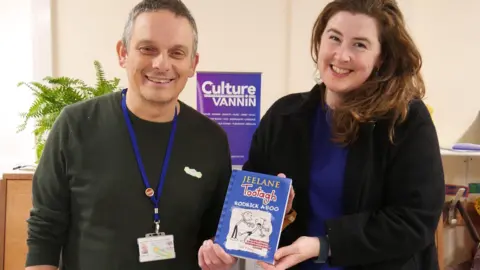 CULTURE VANNIN Adrian Cain, who has short greying hair and is wearing a black jumper, and Ruth Keggin-Gell, who has long brown hair and is wearing a black jacket over a blue top, holding a copy of the book, which has a blue cover with a black and white illustration. There is a purple Culture Vannin banner in the background.