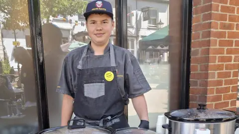 Dexter stood in his all grey chef uniform stood in front of slow cookers on a table with recipe cards, spices, tins and flatbreads on the table.
