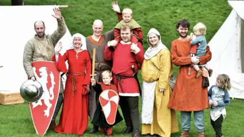 City of Lincoln Council A group of re-enactors at the Lincoln Festival of History in 2024. The picture shows four men, two women, and four young children. They are dressed in historical outfits. Two of the group are carrying shields and one of the boys has a wooden sword. 