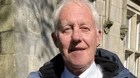 Oldham councillor Graham Sheldon smiles for the camera. He is wearing a white shirt, tie, and a navy blue coat.