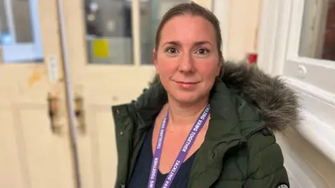 Detective Chief Inspector Lucy Thorne from Wiltshire Police. She has dark hair which is tied back. She is pictured looking directly at the camera and smiling. She is wearing a dark green parka with a fur lined hood and a navy v-neck t-shirt. She also has a purple lanyard.