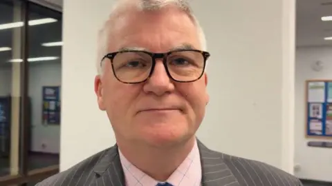Martin Heath/BBC Graeme Coombes with short white hair and black framed glasses, wearing a grey pin-striped jacket, pink shirt and blue tie. He is standing in a council foyer with a white wall behind.