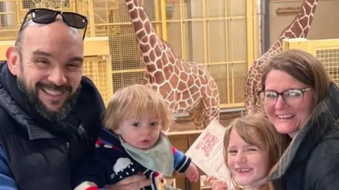 Family photo A family picture at a zoo with giraffes in the background. Gary is bald with a dark beard and moustache, wearing a black puffer coat and holding a toddler with blonde hair, who is wearing a blue cardigan and light green bib. Next to him is a young girl with blonde hair and a tooth gap who is hugging Natalie who has long brown hair and glasses in a black puffer coat. They are all smiling at the camera.