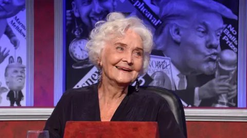 BBC/Hat Trick Woman with grey hair in dark clothing in TV studio smiling at a red desk