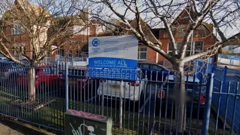 A google streetview image of the Castle Hill Community Primary School in Folkestone, Kent. The school is a red brick building; visible from the outside is a blue fence and a sign welcoming visitors to the school. 