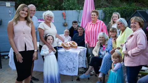 A photo of me and my family celebrating my auntie's 80th birthday in the garden. There are 14 of us, including three children, all smiling at the camera. My auntie is seated in the middle, wearing a pink birthday badge. On the table are two cakes shaped as the number 80. There are birthday banners on the wall in the background. 