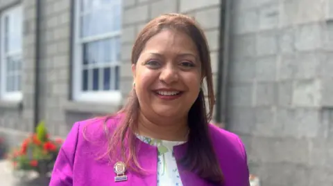 Munazza Malik wearing a bright purple jacket with a floral white t-shirt beneath her jacket. She has long brown hair and has been photographed in front of a grey stone wall.
