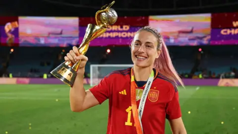 Alexia Putellas, a Spanish footballer wearing a red jersey, holding a golden trophy