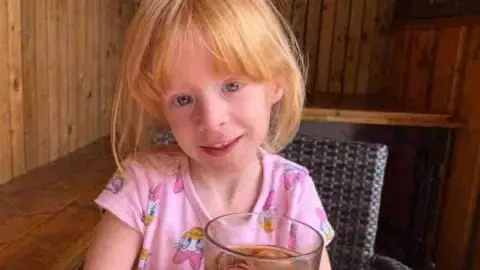 Handout Holly Prince, who has ginger hair and is wearing a pink Disney themed top, smiles at the camera while sitting at a table in a restaurant or cafe with a glass of cola in front of her. 