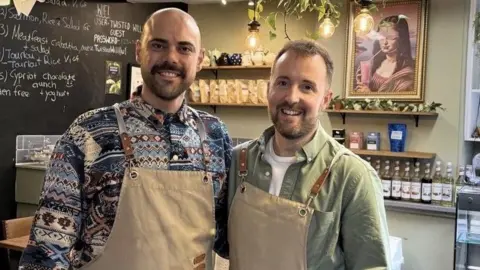 Constantinos Theophanous and Scott Thain stand together in a cafe. They are wearing brown aprons and are both smiling. There is a black board with drinks and food listed on it and to the right of them there are several syrups and coffees and teas. Constantinos has no hair and is wearing a patterned blue shirt while Scott has short brown hair and is wearing a green shirt.