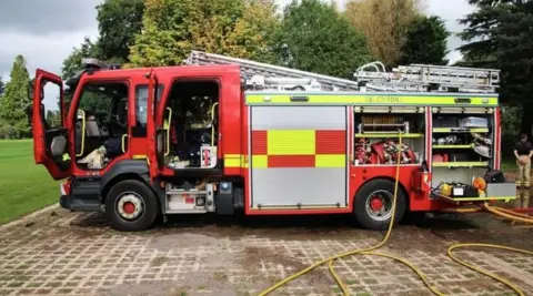 A fire engine is parked on a hard-standing next to a field. It has its doors open and one of its hoses is lying on the ground after being unravelled. Trees can be seen in the background.