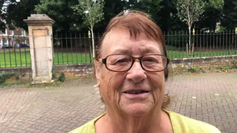 Karen Youngman standing in front of the fences of the Wisbech War Memorial Garden. She is wearing glasses and a yellow top.