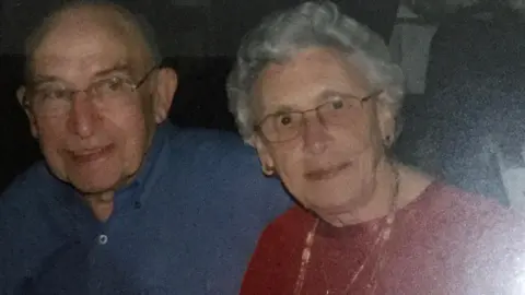 Joe and Rose Slyper sit closely side by side indoors, both wearing glasses and smiling softly at the camera. He wears a blue shirt; she wears a red top with a beaded necklace and has short curled grey hair