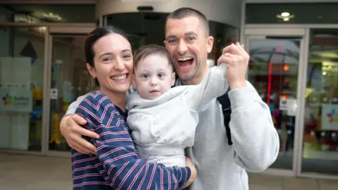 PA Media A slim woman with dark hair tied back, wearing a navy blue and red stripe top and a dark haired man in a grey hoodie. They stand together, holding a dark haired male toddler, both smiling. Callum holds up the boy's arm as though in celebration.
