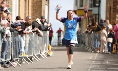 Apryl Hammett Apryl holds her arms high as she nears the finish of a recent marathon. She is wearing a light blue, purple and yellow vest and she's cheered across the line by spectators who are applauding outside a barrier at the finish.