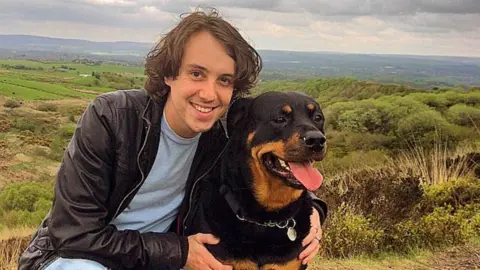 Oliver Robinson with medium length brown hair, wearing a dark jacket and light blue tshirt smiles at the camera with his arms around a black and tan rottweiler dog with countryside and fields in the background