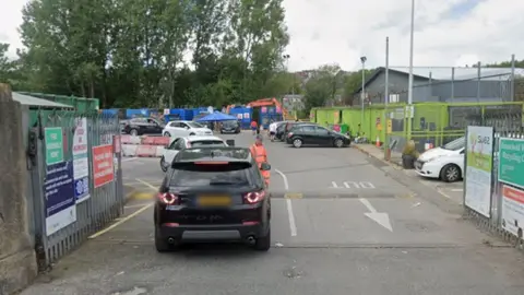 Google Entrance to the George Street West site, showing a black car driving in, with multiple cars parked by an array of blue skips