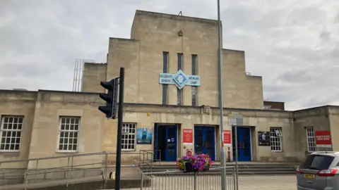 Alex Pope/BBC A large stone building with three sets of blue doors at its front. At the top of the building is a blue sign that reads "Mounts Baths Health Suite". 