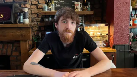 A man sits at a wooden bar inside a traditional pub, with a drinks menu, brick walls and shelves of bottles in the background.