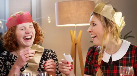 Getty Images Two women wearing Christmas hats are sharing a joke from a pulled cracker.