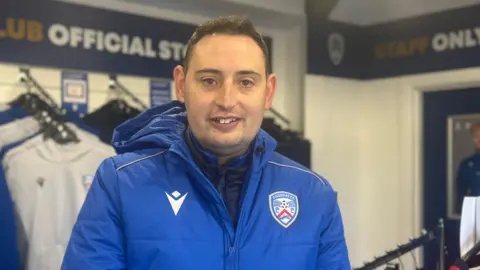 Scott Smyth is a man with dark short hair. He is standing in the shop with jumpers and hoodies behind him, smiling at the camera.
He is wearing a bright blue sports coat with the Coleraine FC emblem on the right hand side.