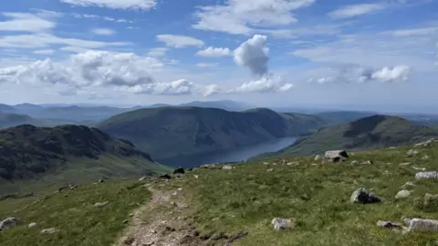 Butterfly Conservation Mountain range with waterbody going through it. There are clouds in the blue sky. It is sunny. There are rocks on the grass on the clifftops. 