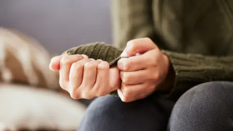 Getty Images A woman sits on a sofa. She is pulling on a sleeve of her green jumper in a sign of feeling anxious.