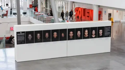 Nosa Malcolm/OG A giant wide white plinth with 10 headshot images is on display inside a building with a mezzanine floor visible in the background with people walking past.