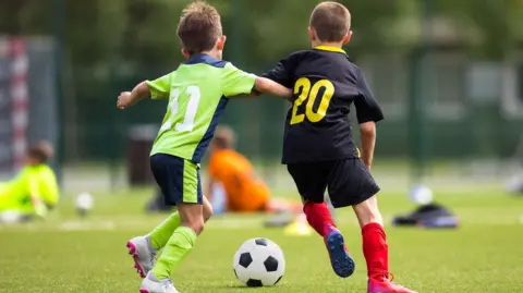 Two young boys wearing football kits playing football. One in a green kit has his arm outstretching touching the other boy who is in a black kit. There is a black and white ball in front of them.
